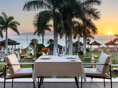 Tafel met twee stoelen en wijnglazen bij palmbomen met zeezicht bij zonsondergang.