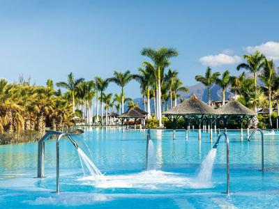 Large pool with water jet and palm trees in tropical resort under clear sky.