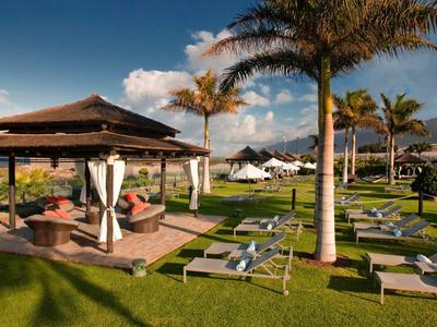Groene tuin met ligstoelen, paviljoenen en palmbomen aan zee bij heldere lucht.