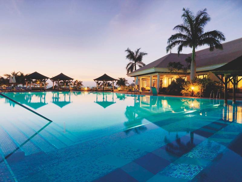 Verlicht zwembad met palmbomen en strandparasols bij zonsondergang naast een resort.