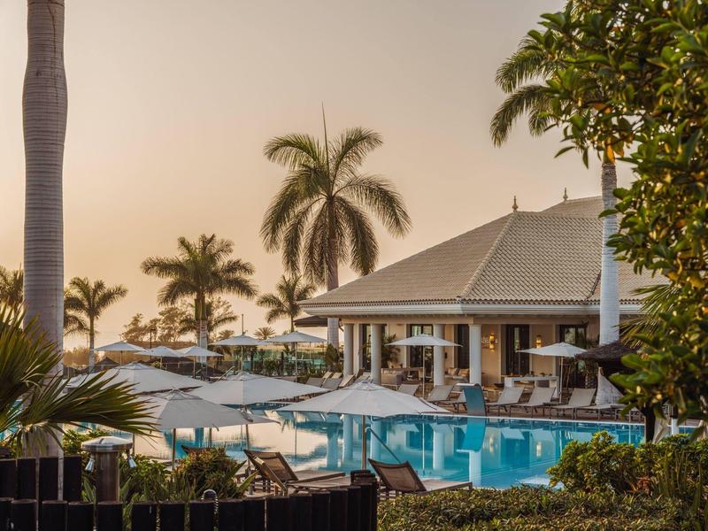 Hotelzwembad met palmbomen en parasols bij zonsondergang.