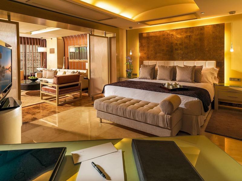 Groot hotelzwembad met ligstoelen en palmbomen voor een gebouw met een licht dak.