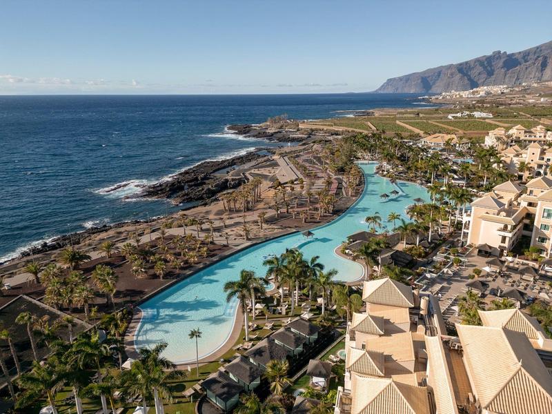 Luchtfoto van een resort met groot zwembad, palmbomen en uitzicht op zee langs een rotskust.
