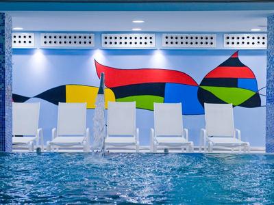 Innen-Schwimmbad mit blauem Wasser, weißen Liegestühlen und einer farbenfrohen Wandmalerei.