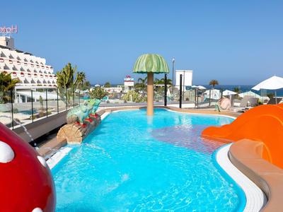 Bunter Kinder-Pool mit Pilzwasserfontäne, Rutschen und blühender Dachterrasse bei klarem Himmel.