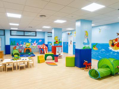 Heller Kindergartenraum mit bunten Möbeln, Spielzeug, Wandbildern und Teppichen auf hellem Boden.