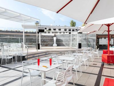 Area esterna moderna con tavoli bianchi, sedie e grandi ombrelloni in una giornata di sole.