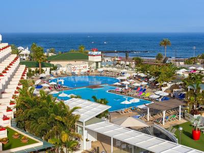 Großer Hotelpool mit Liegestühlen, Palmen und Blick auf das blaue Meer im sonnigen Urlaubsort.