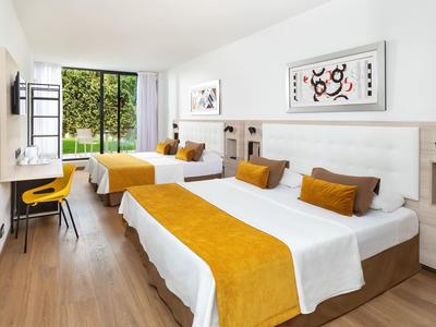 Hell eingerichtetes Hotelzimmer mit zwei großen Betten, gelben Akzenten und Blick auf Garten.