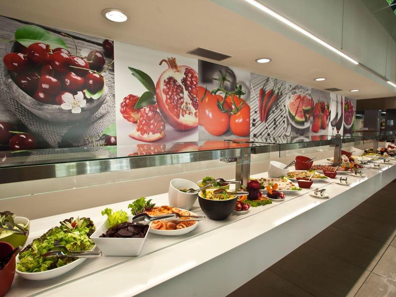 Langes Buffet mit frischem Salat und Gemüse in Schüsseln, dekoriert mit großformatigen Obstfotos an der Wand.