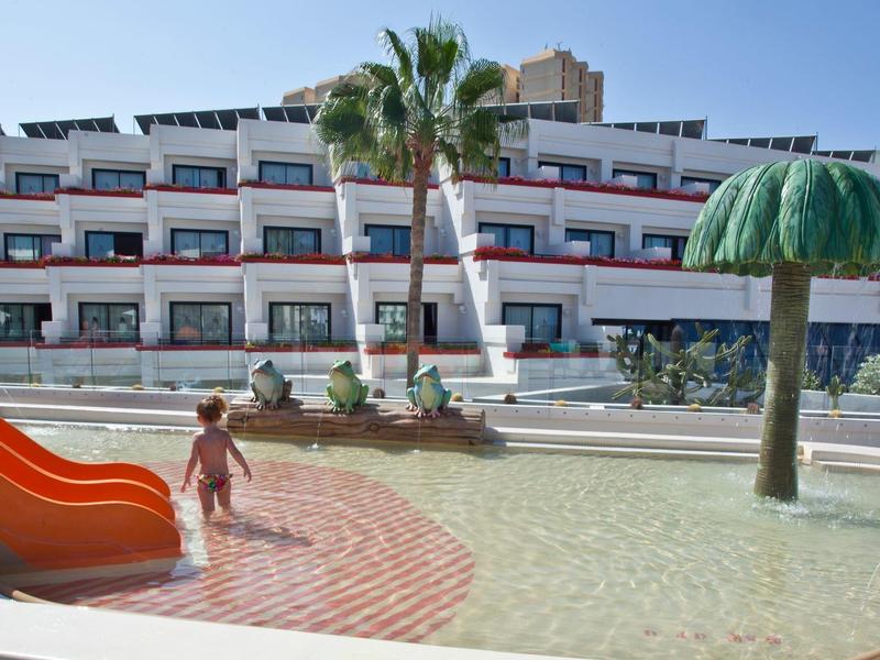 Kind spielt im flachen Wasser eines Kinderpools mit Rutsche neben Palmen und einem Hotelgebäude.