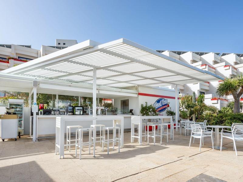 Moderner Außenbarbereich mit weißen Möbeln und Sonnendach vor mehrstöckigem Hotel.