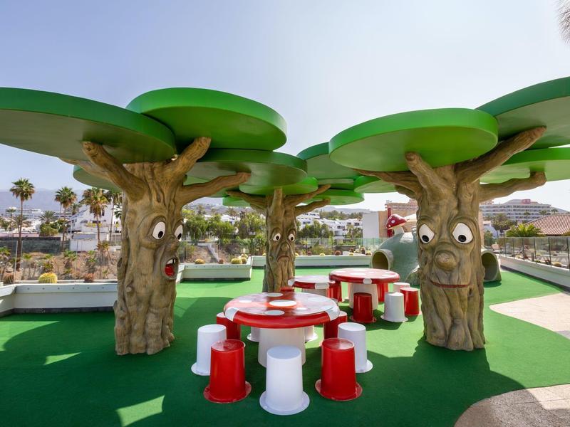 Bunter Kinderspielplatz mit Baum-Skulpturen und rot-weißen Sitzgelegenheiten unter blauem Himmel.