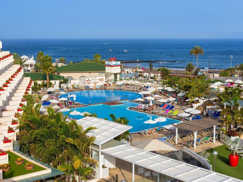 Großer Hotelpool mit Liegestühlen, Palmen und Blick auf das blaue Meer im sonnigen Urlaubsort.