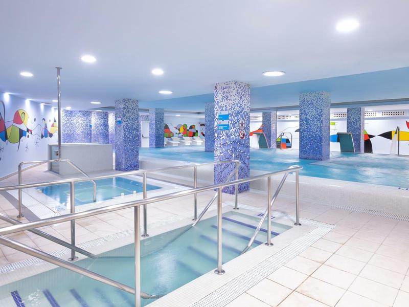 Helles Hallenbad mit Treppen, Handläufen, bunten Wandgemälden und ruhigem Wasser.