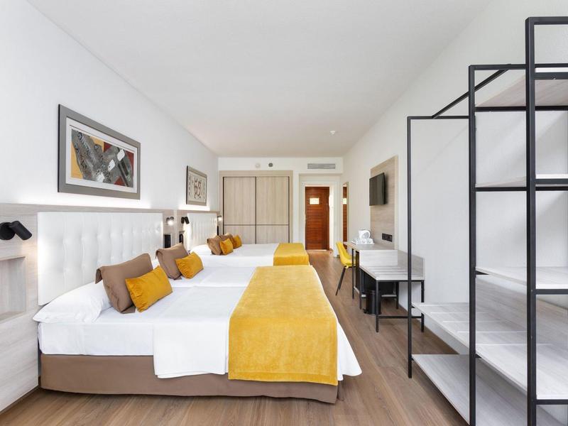 Helles, modernes Hotelzimmer mit zwei Doppelbetten, weißen Wänden, gelben Akzenten und offenem Regal.