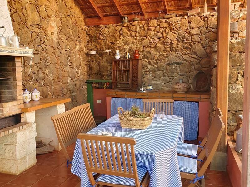 Pohodlná jídelní část s dřevěnými židlemi, stolem s modrým ubrusem a kamennou zdí pod střechou.