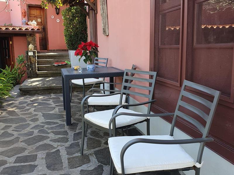 Dvě židle s bílými polštáři a stůl s červenou květinovou dekorací na kamenné terase před růžovou budovou.