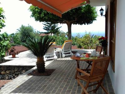 Terrasse avec chaises et tables, auvent orange et quelques plantes à côté d'une maison.