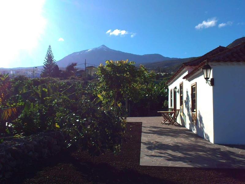 Vue ensoleillée d'un bâtiment blanc à côté de plantes vertes avec des montagnes en arrière-plan.