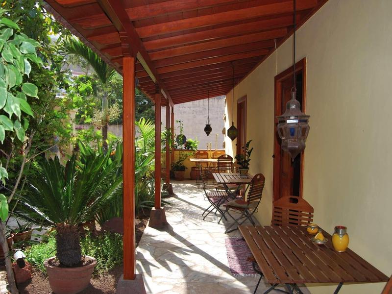 Terrazza coperta con tavoli in legno e piante lungo il lato di un edificio giallo.