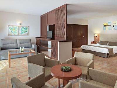 Camera d'hotel luminosa con letto, divano, zona pranzo e divisorio in legno con grandi finestre.