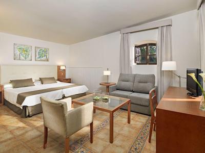 Camera d'hotel luminosamente arredata con letto matrimoniale, divano, poltrone e TV.