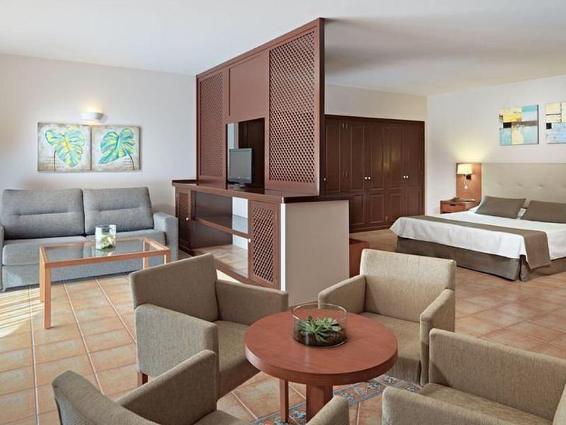 Camera d'hotel luminosa con letto, divano, zona pranzo e divisorio in legno con grandi finestre.