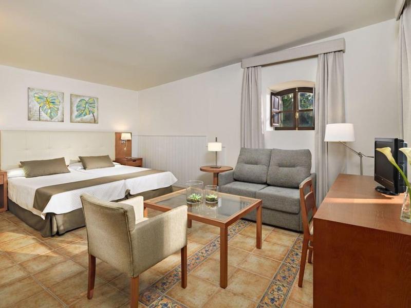 Camera d'hotel luminosamente arredata con letto matrimoniale, divano, poltrone e TV.