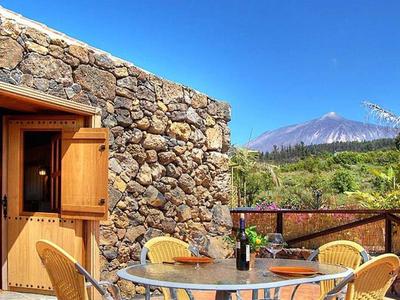 Steinmauer mit Holztür, davor Tisch mit Stühlen, Weinflasche und Gläser, im Hintergrund Berg und blauer Himmel.