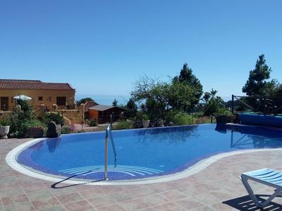 Piscina rotonda all'aperto con corrimano, circondata da piastrelle di pietra e lettini, accanto a edifici e alberi sotto un cielo blu.