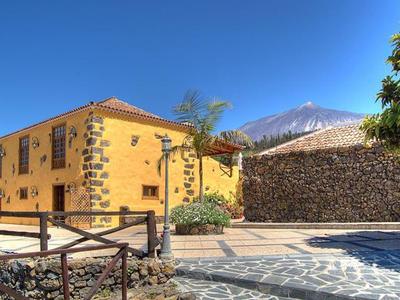 Gelbes Haus neben gemauertem Zaun mit Palmen, blauer Himmel und schneebedecktem Berg im Hintergrund.