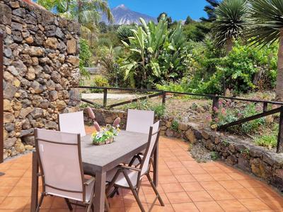 Terrasse mit braunen Kacheln, Holztisch, vier Stühlen, blühenden Pflanzen und Steinmauer im Garten.