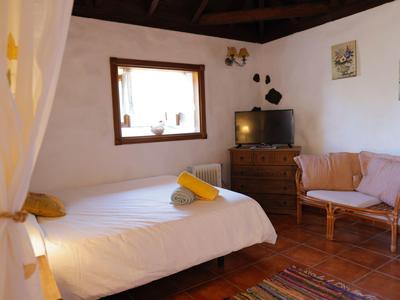 Gemütliches Schlafzimmer mit weißem Bett, Holzfenster, Fernseher, Kommode und Sofa in warmen Tönen.