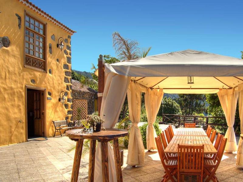 Sonnige Terrasse mit Holztisch, Stühlen und weißem Pavillon neben gelbem Steinhaus.