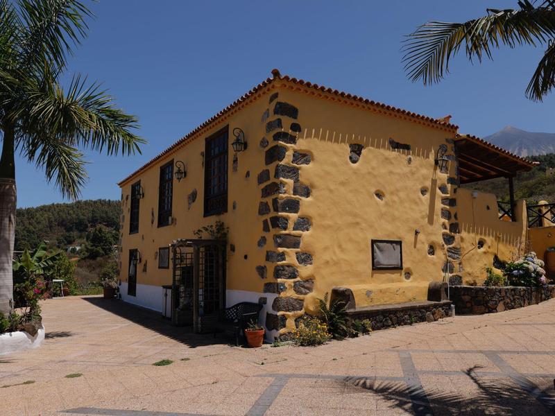 Gelbes, zweistöckiges Haus mit dunklen Steinecken, umgeben von Palmen und klarem Himmel.