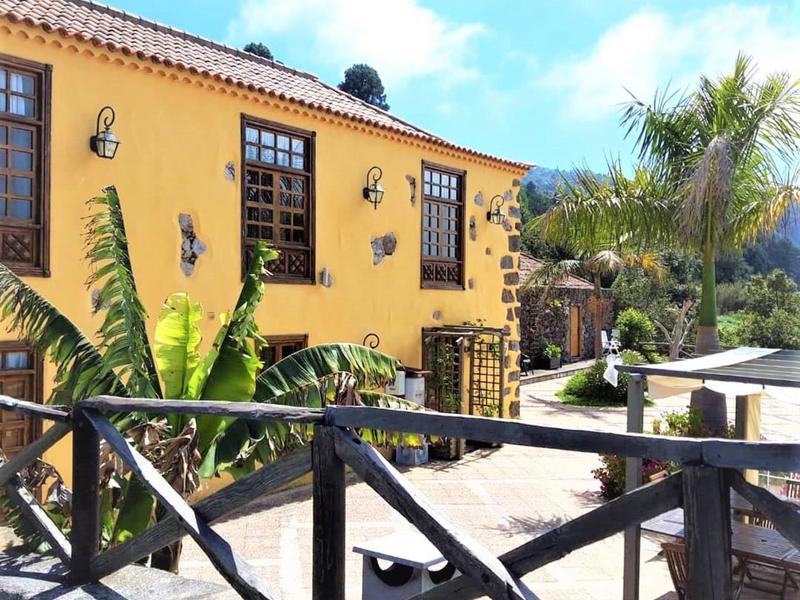 Haus mit gelb-orange Fassade, Pflanzen davor, Holzzaun und blauer Himmel mit Wolken.
