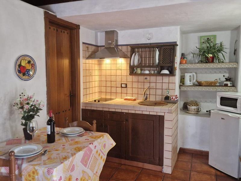 Kleine Küche mit Holzmöbeln, Fliesenrückwand, Esstisch mit Blumen und Flaschen, weiße Wände.