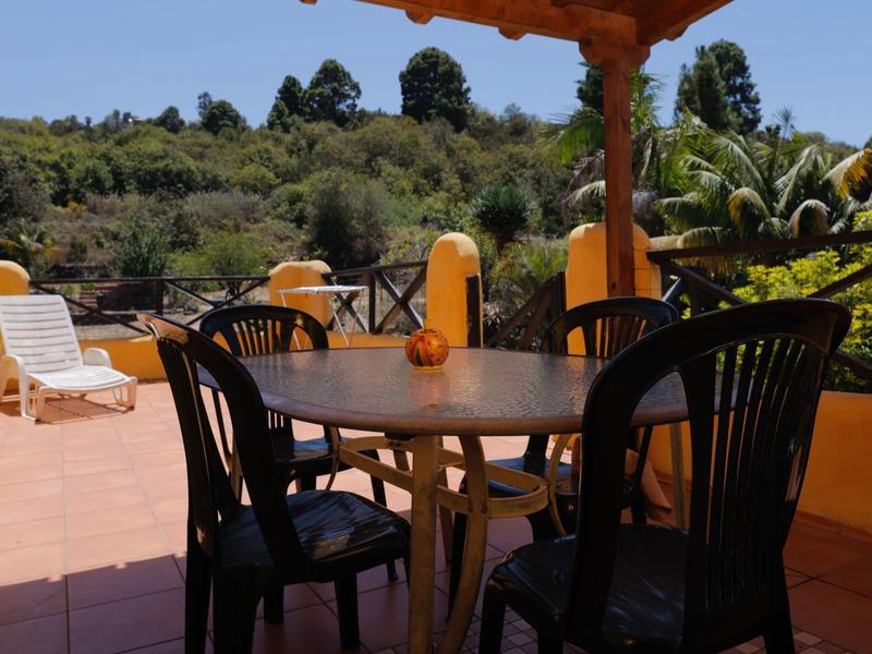 Terrasse mit rundem Tisch, vier schwarzen Stühlen und Sonnenschirm, umgeben von gelben Wänden.