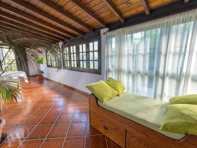 Veranda luminosa con soffitto in legno, cuscino verde e grandi finestre con tende bianche.