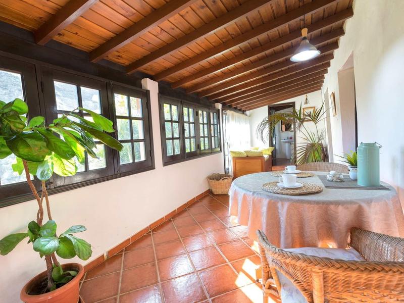 Veranda luminosa con soffitto a travi in legno, piante e tavolo apparecchiato con sedie in vimini.