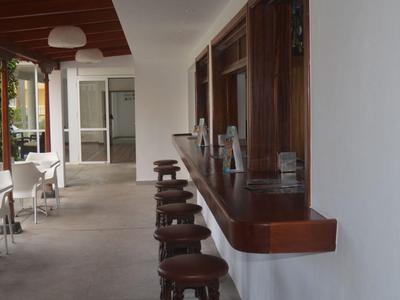 Überdachte Terrasse mit Holztresen, Barhockern und weißen Tischen und Stühlen neben einer Hecke.