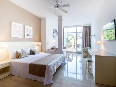 Bright hotel room with double bed, desk, chairs, and wall-mounted TV.