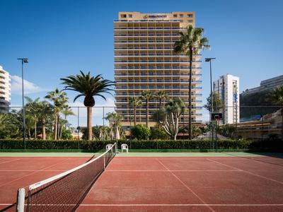 Tennis court in front of a tall hotel building with palm trees and clear sky.
