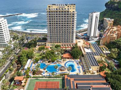 Vista de un hotel con piscina, pista de tenis y mar al fondo.