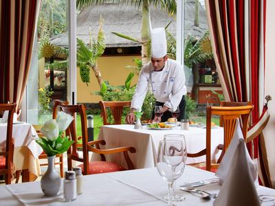 Waiter in white uniform sets a table in a restaurant overlooking a garden.