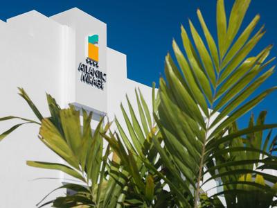 Bílá budova hotelu s logem na pozadí jasné modré oblohy a zelené palmové listy v popředí.