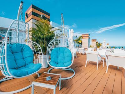 Moderní lounge s modrými závěsnými křesly a bílými židlemi na dřevěné terase pod modrou oblohou.