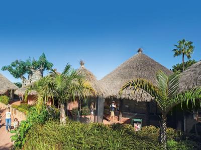 Vue d'un complexe tropical avec des palmiers et des huttes au toit de chaume sous un ciel bleu.