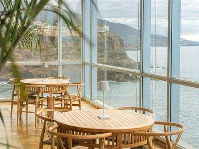 Dřevěné stoly a židle v prosklené místnosti s výhledem na moře a skály.